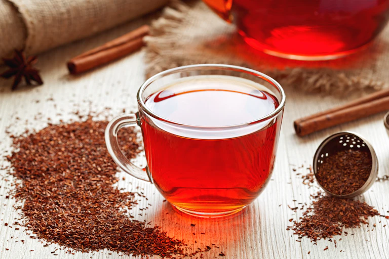 A cup of rooibos tea on a table, surrounded by loose tea, a teapot and spices