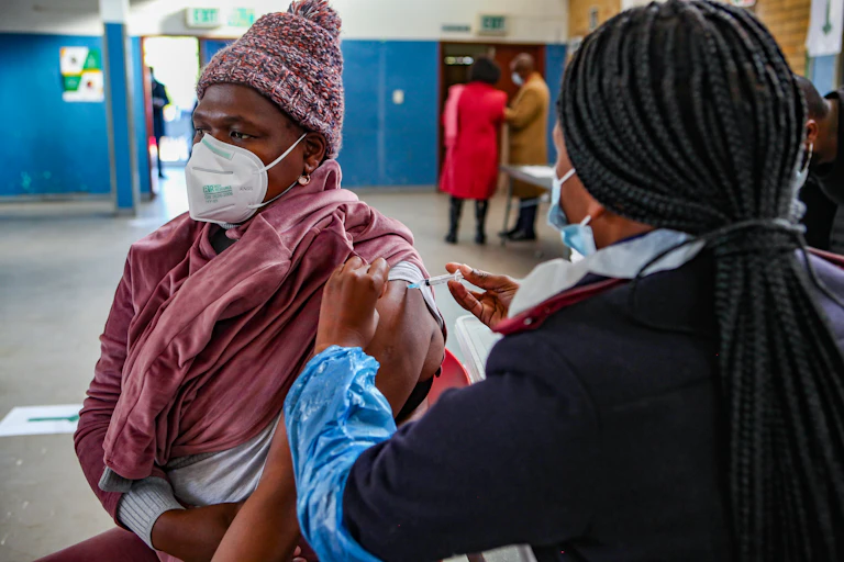 Woman wearing face mask, one sleeve rolled up, with another woman in nurse's uniform injecting her exposed arm