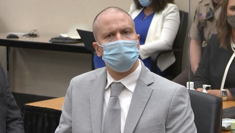 A white man wearing a gray suit and a blue mask listens to an unseen speaker