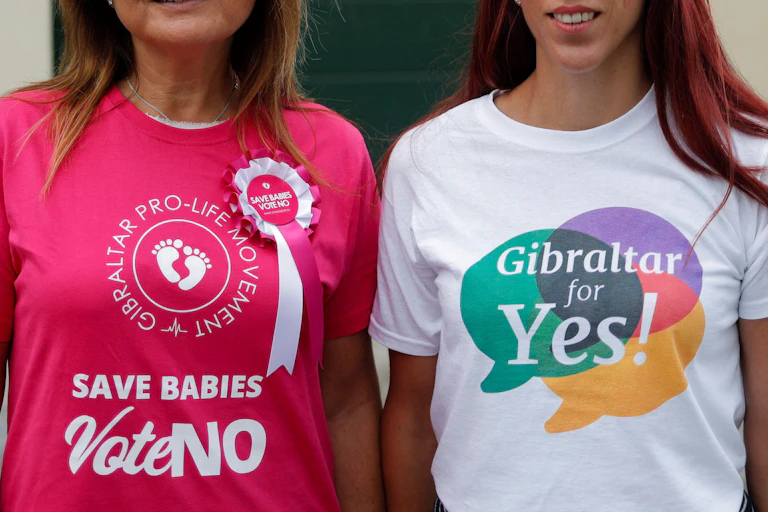 Two women standing side by side, one with a t-shirt reading "save babies vote no," the other with a shirt reading "Gibraltar for yes"