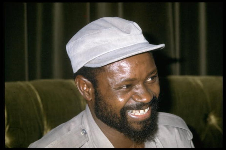 A bearded man wearing a military cap and shirt smiles heartily.