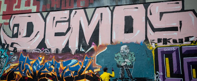 A man stands infront of a mural depicting Bernie Sanders. The word demos is written above.