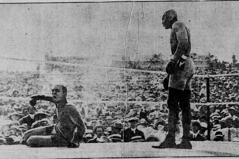 A Black boxer at right stands over a fallen white boxer at left