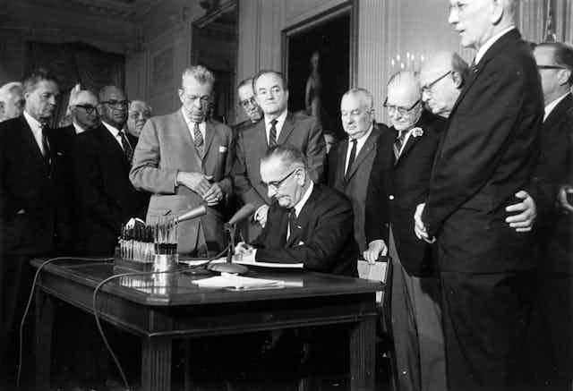 President Lyndon Johnson at a desk, surrounded by white lawmakers, as he signed the 1964 Civil Rights Act with multiple pens.
