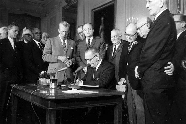 President Lyndon Johnson at a desk, surrounded by white lawmakers, as he signed the 1964 Civil Rights Act with multiple pens.
