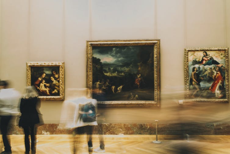 People walk past paintings in a museum.