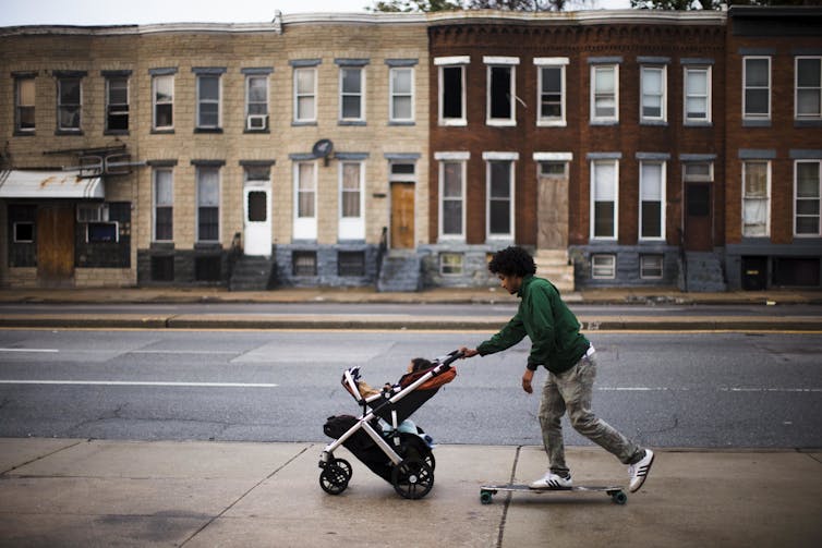 Um jovem negro de skate empurra o filho em um carrinho de bebê em uma calçada, passando por prédios destruídos em Baltimore.