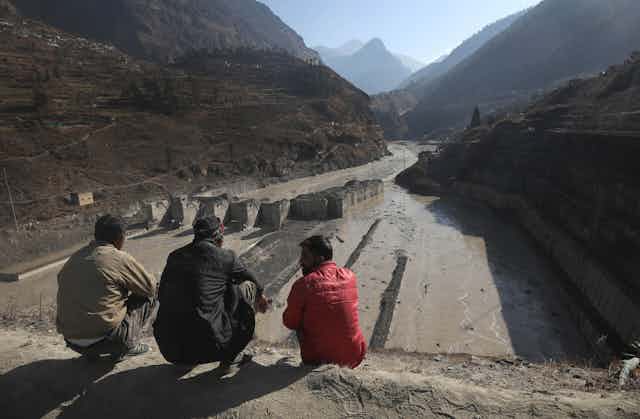 Three men sit on a rise overlooking a damaged hydropower project. 