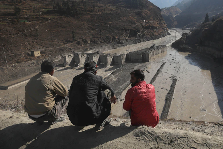 Three men sit on a rise overlooking a damaged hydropower project.