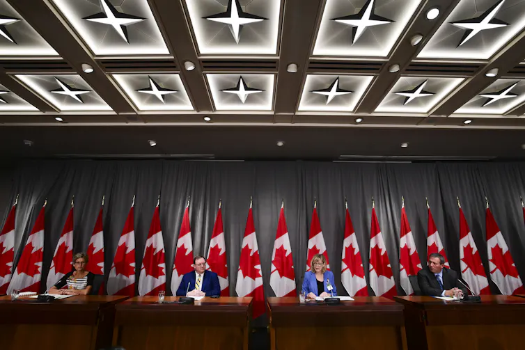 four government officials seated in front of a row of flags