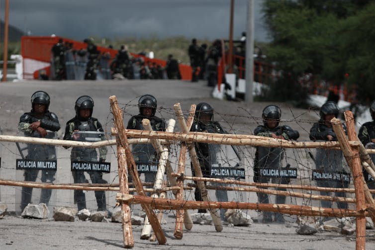Soldiers in riot gear stand behind a fence wrapped with barbed wire.