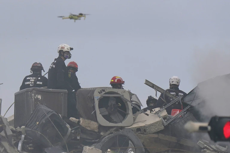 a drone flies above five people in helmets standing on a pile of metal and concrete rubble