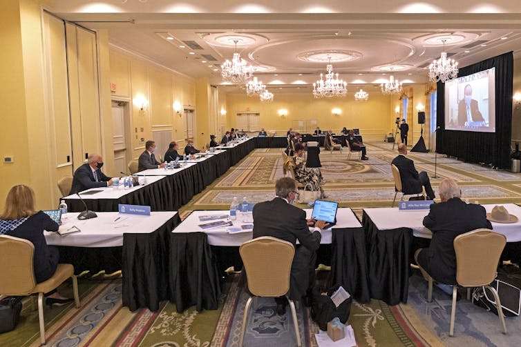 Many people meeting around a U-shaped black-skirted table, looking at a large screen