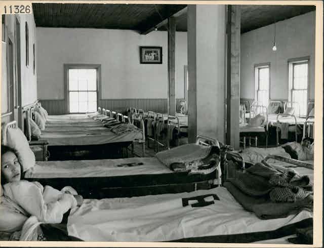 Deux filles sont couchées sur des lits de dortoir dans un pensionnat autochtone.