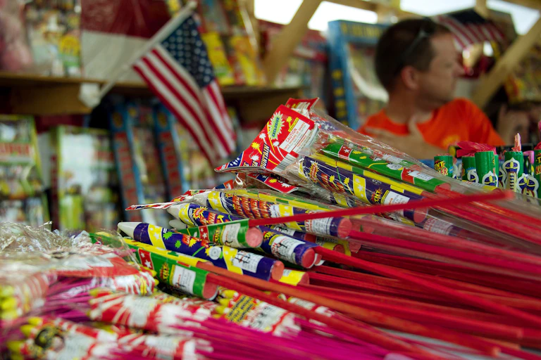 A man sits near a stack of bottle rockets with a flag in the background.