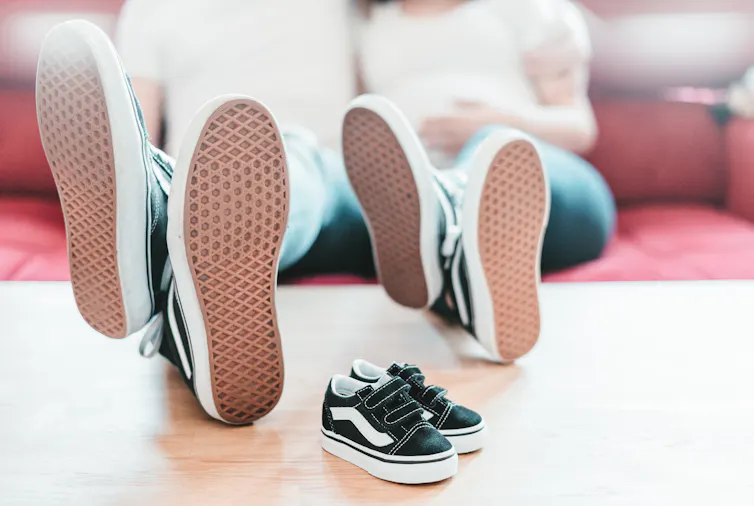 A couple sits with their feet up on the table wearing matching shoes, a pair of kids sneakers is on the table in front of them.