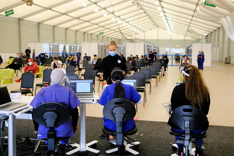 Workers at a mass vaccination hub sit in chairs with their back to the camera.