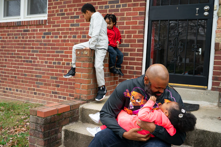 Man hugs little girl as two boys play behind them