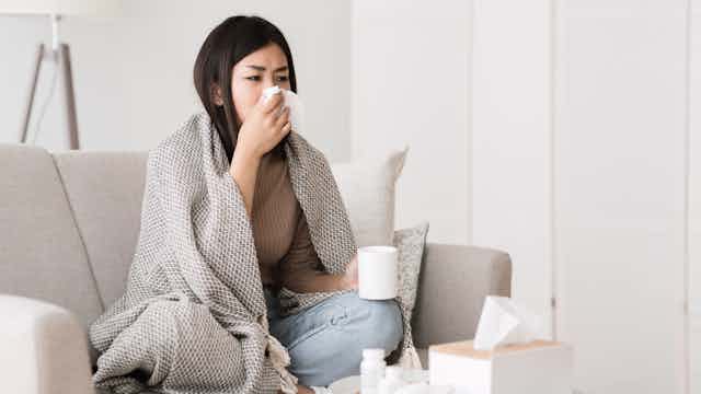 A young woman sits on the couch wrapped in a blanket, blowing her nose.