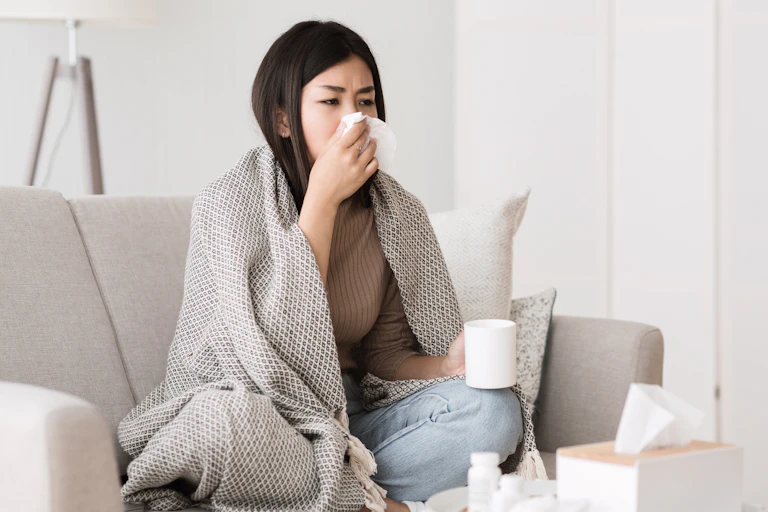 A young woman sits on the couch wrapped in a blanket, blowing her nose.