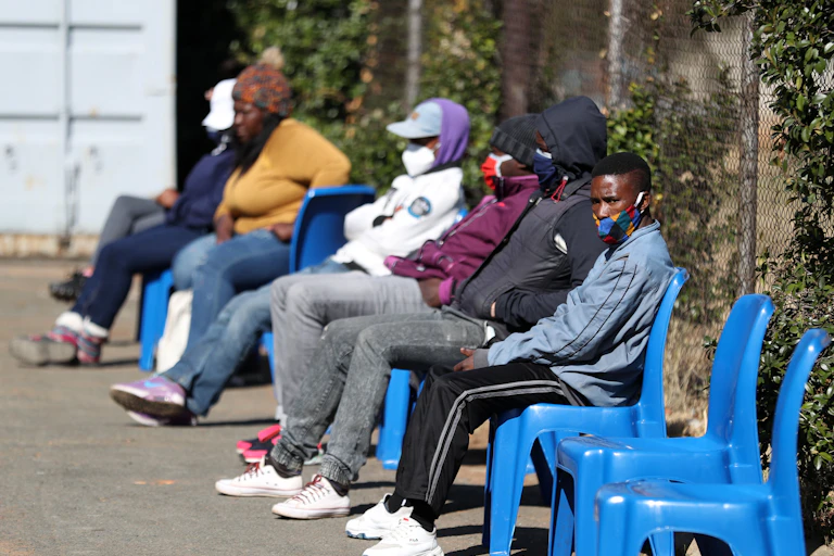 Six people, wearing face masks, sit in a row of plastic chairs outdoors