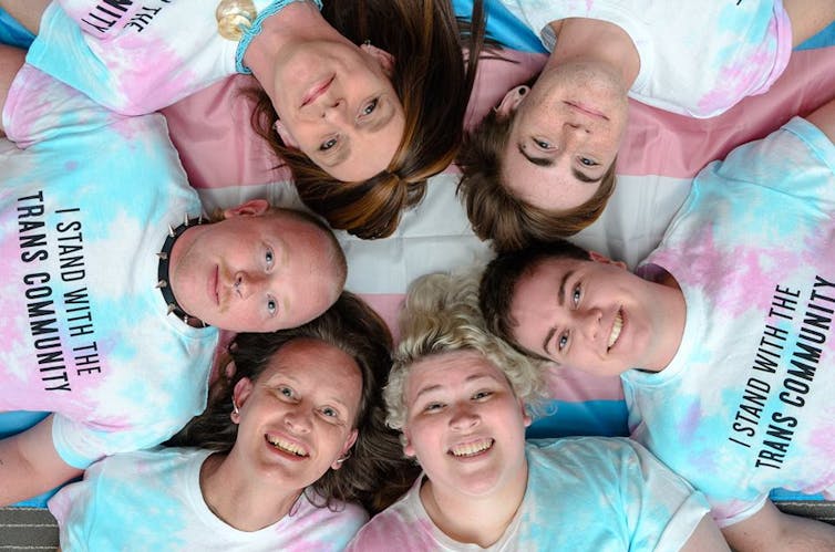 Six people laying on the ground in a circle with tie dyed t-shirts that say 'I stand with the trans community.'