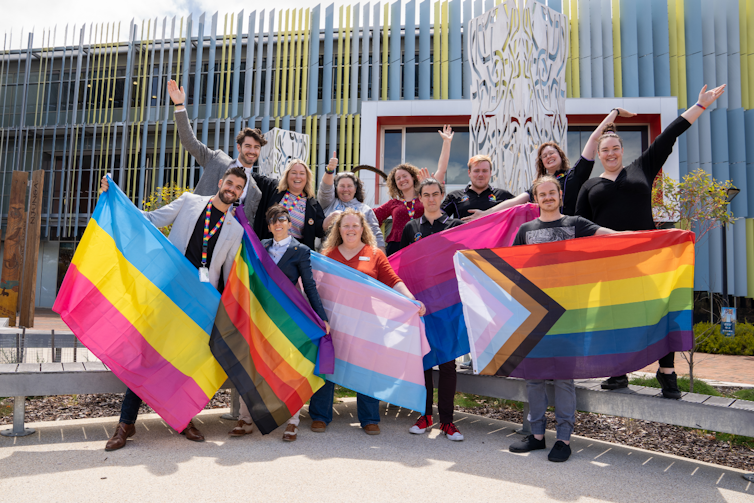ECU staff and students hold up the pansexual flag, rainbow flag, trans flag, bisexual flag and progress pride flag with their hands waving in the air and smiling.