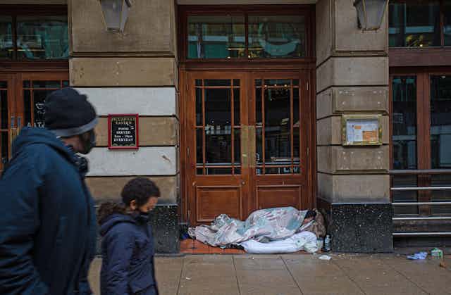 A mother and son wearing masks walk past a homeless person sleeping in front of a doorway