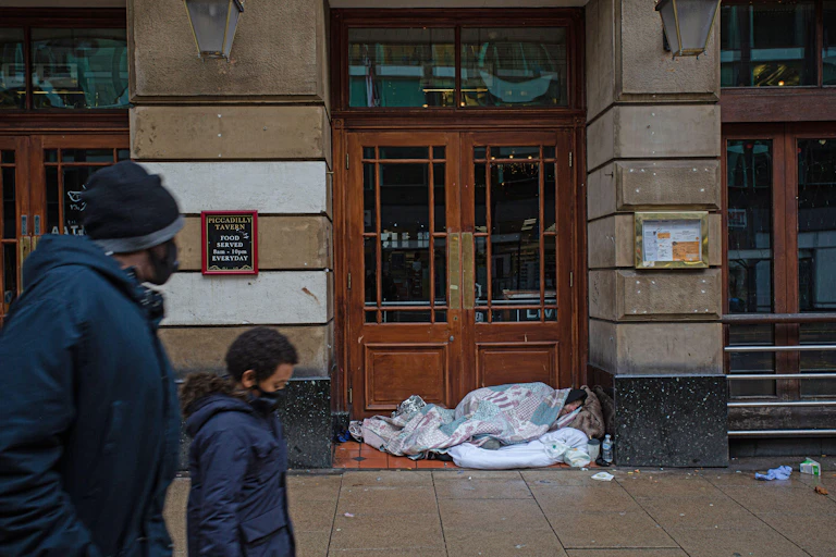A mother and son wearing masks walk past a homeless person sleeping in front of a doorway