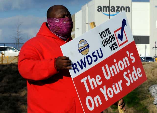 A Black man in a red coat and a purple face mask holds a pro-union sign outside of an amazon facility 
