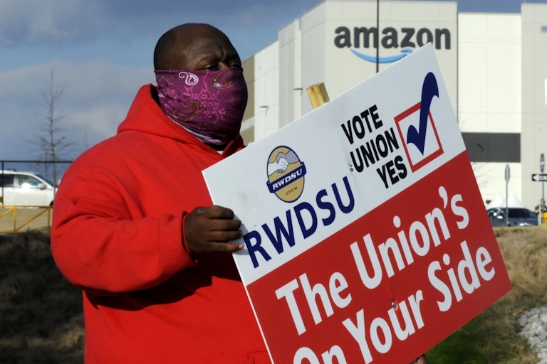 A Black man in a red coat and a purple face mask holds a pro-union sign outside of an amazon facility