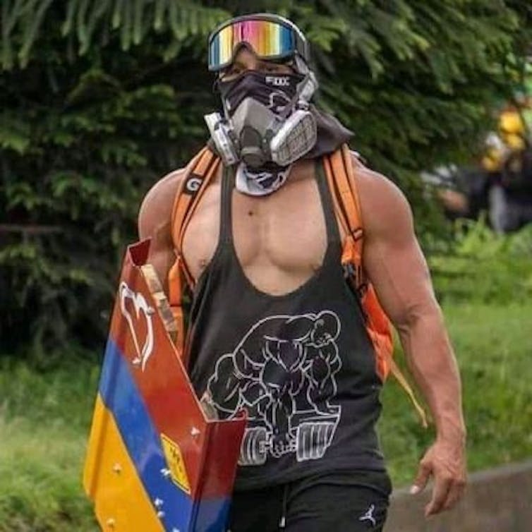 A muscular man in a gas mask, ski goggles, and a tank top, holding a metal shield painted like the Colombian flag
