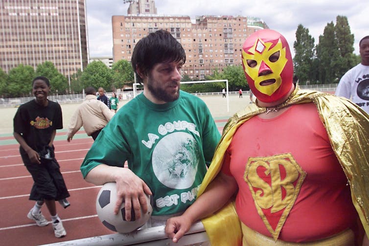 A man holding a soccer ball stands next to a man wearing a red full-face mask with a cape and an 'SB' emblem on his shirt.