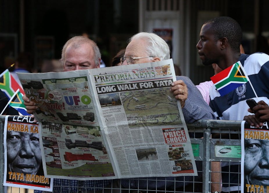 A young man holding a miniature South African flag in his left hand, reads a newspaper over the shoulders of two balding, elderly men.
