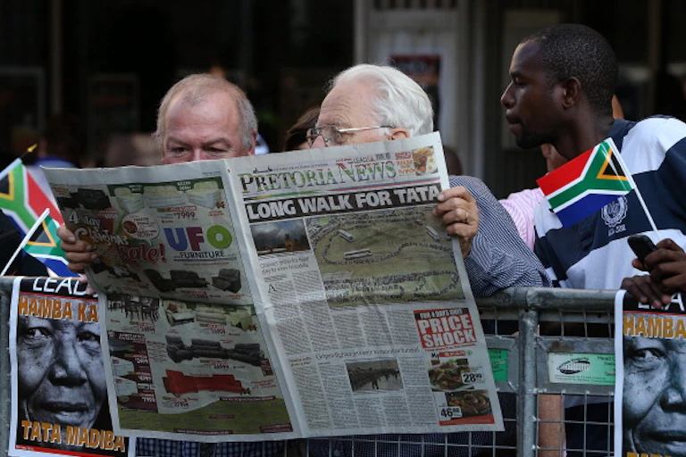 A young man holding a miniature South African flag in his left hand, reads a newspaper over the shoulders of two balding, elderly men.
