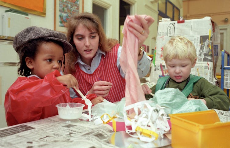 A pre-school teacher holds up a piece of fabric during a crafting session with two boys in aprons