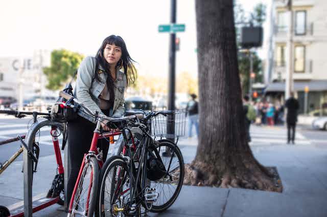 On a busy city sidewalk, one Latina leans over to lock her bike before heading into work. She's dressed casually with denim jacket and black jeans, a shoulder bag hangs from one shoulder. There are two other bikes in the rack already. A slight breeze lifts her long dark hair and we see dozens of pedestrians in soft focus in the background waiting at a crosswalk. A very old tree trunk stands tall and strong next to the bike rack, roots lifting the pavement in this place where nature and city come together in balance.