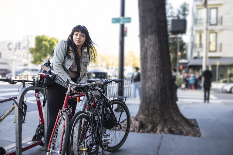 On a busy city sidewalk, one Latina leans over to lock her bike before heading into work. She's dressed casually with denim jacket and black jeans, a shoulder bag hangs from one shoulder. There are two other bikes in the rack already. A slight breeze lifts her long dark hair and we see dozens of pedestrians in soft focus in the background waiting at a crosswalk. A very old tree trunk stands tall and strong next to the bike rack, roots lifting the pavement in this place where nature and city come together in balance.