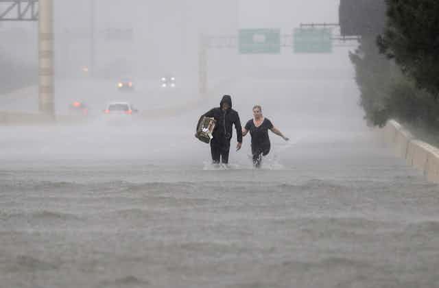 A man and woman walk through knee-high water on an highway in the rain.