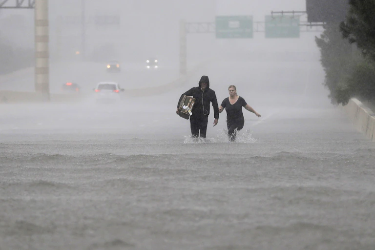 A man and woman walk through knee-high water on an highway in the rain.