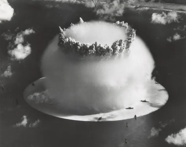 A rapidly expanding cloud of spray from an explosion (nuclear) detonated underwater, framed by small clouds in the air and small black dots around it on the ocean surface which were large naval ships.