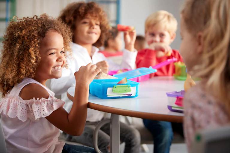 Four young children sit around a table eating from packed lunch boxes