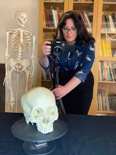 A researcher holding a camera takes a photo of a skull — a skeleton and a bookcase in the background