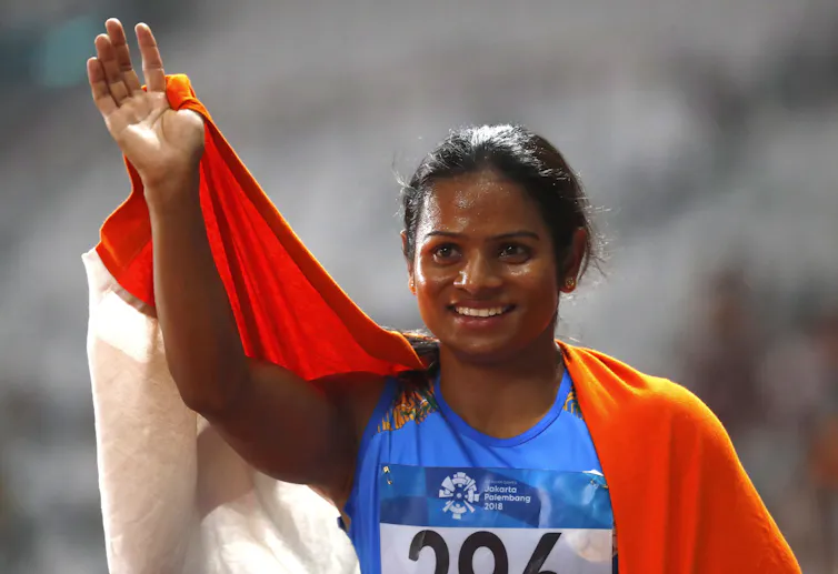 Athlete, with an Indian flag draped around her shoulders, raises one arm to wave at the crowd.