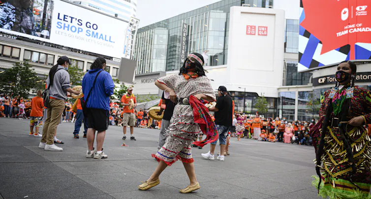 Two jingle dancers dance at a public square under advertisemennts and an ad for Team Canada.