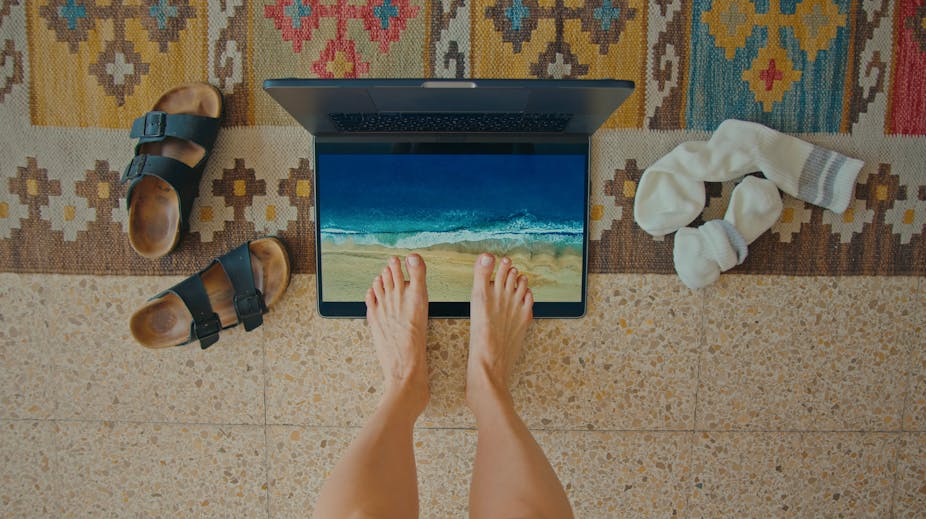 Feet standing on laptop screen showing beach scene with shoes and socks discarded on the floor on either side