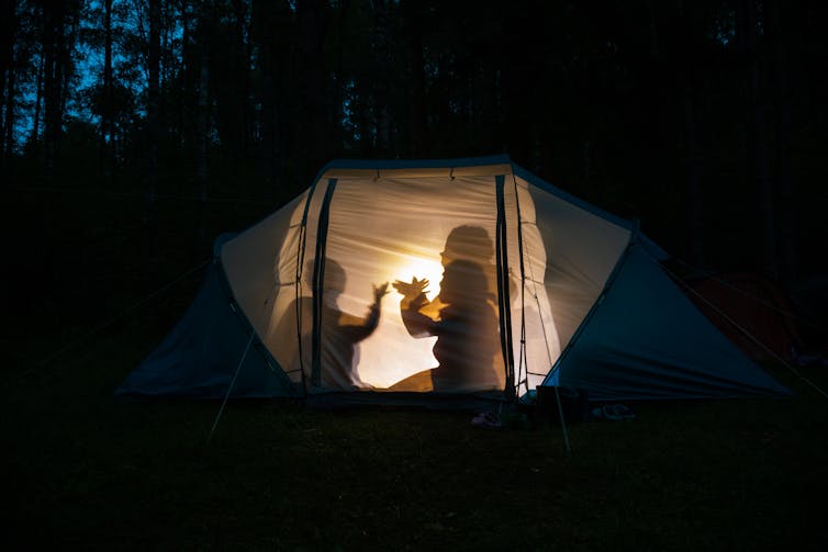 Silhouettes of people making shapes by a light in a tent at night