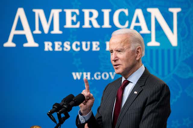 President Biden speaking behind a microphone and in front of a sign that says "American Rescue Plan."