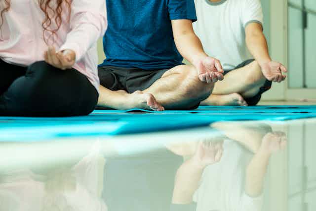 Three people sit on mats with their legs crossed in a meditation pose.