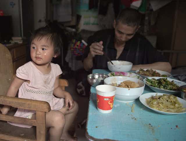 A Chinese toddler eats a big meal with her father in a cramped home.